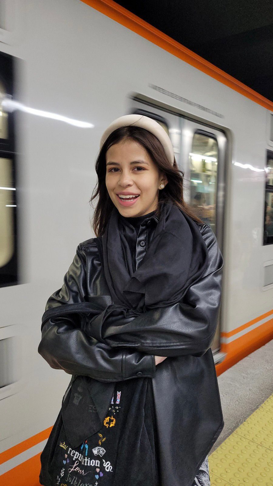 Sofía en el andén del metro, con chaqueta de cuero y diadema, mientras pasa un tren